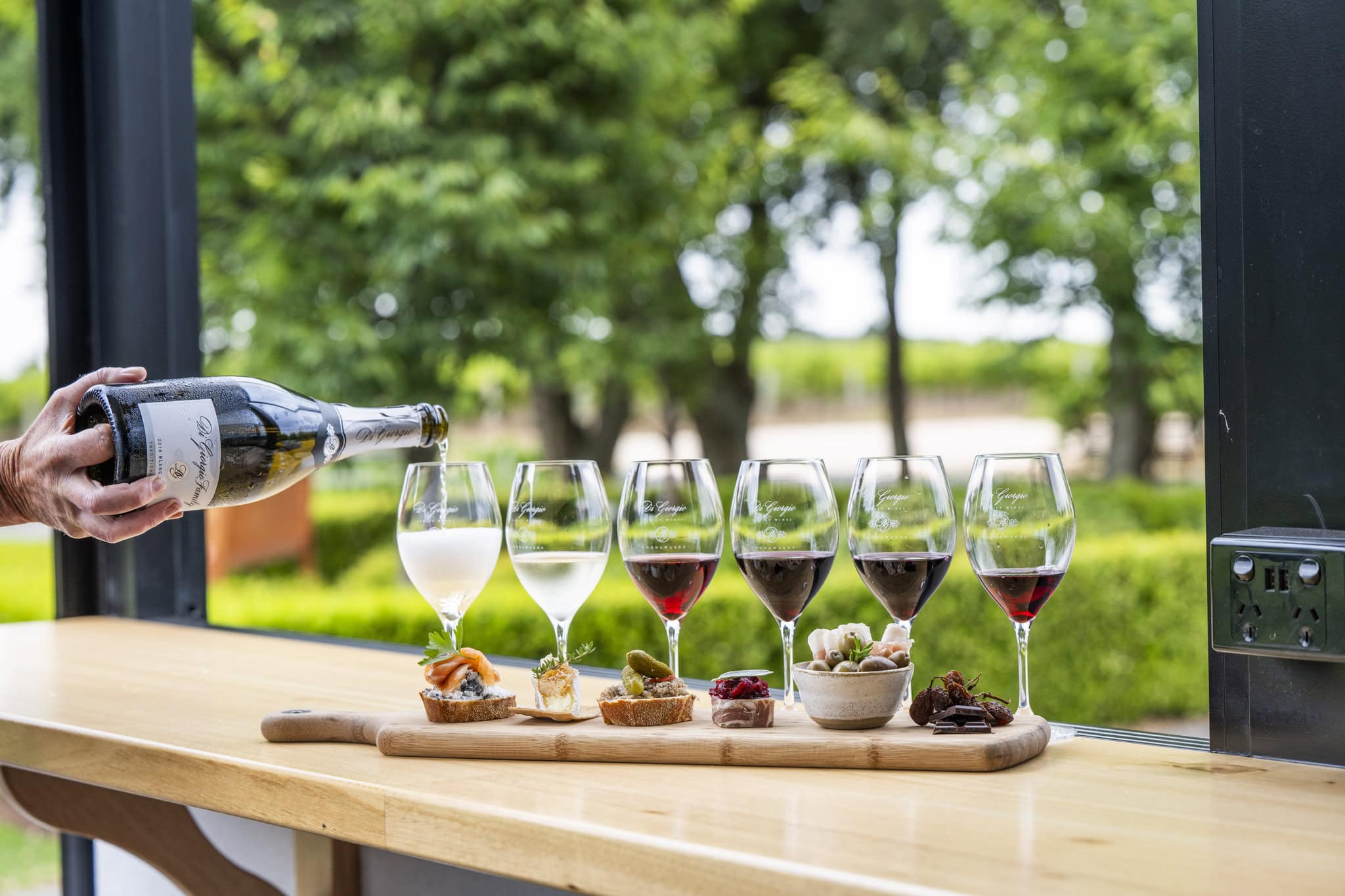 A selection of wine glasses lined up on a bench table overlooking a lush garden. A selection of canape foods sit in front of the wine glasses. Someone pours a sparkling wine into one of the glasses.