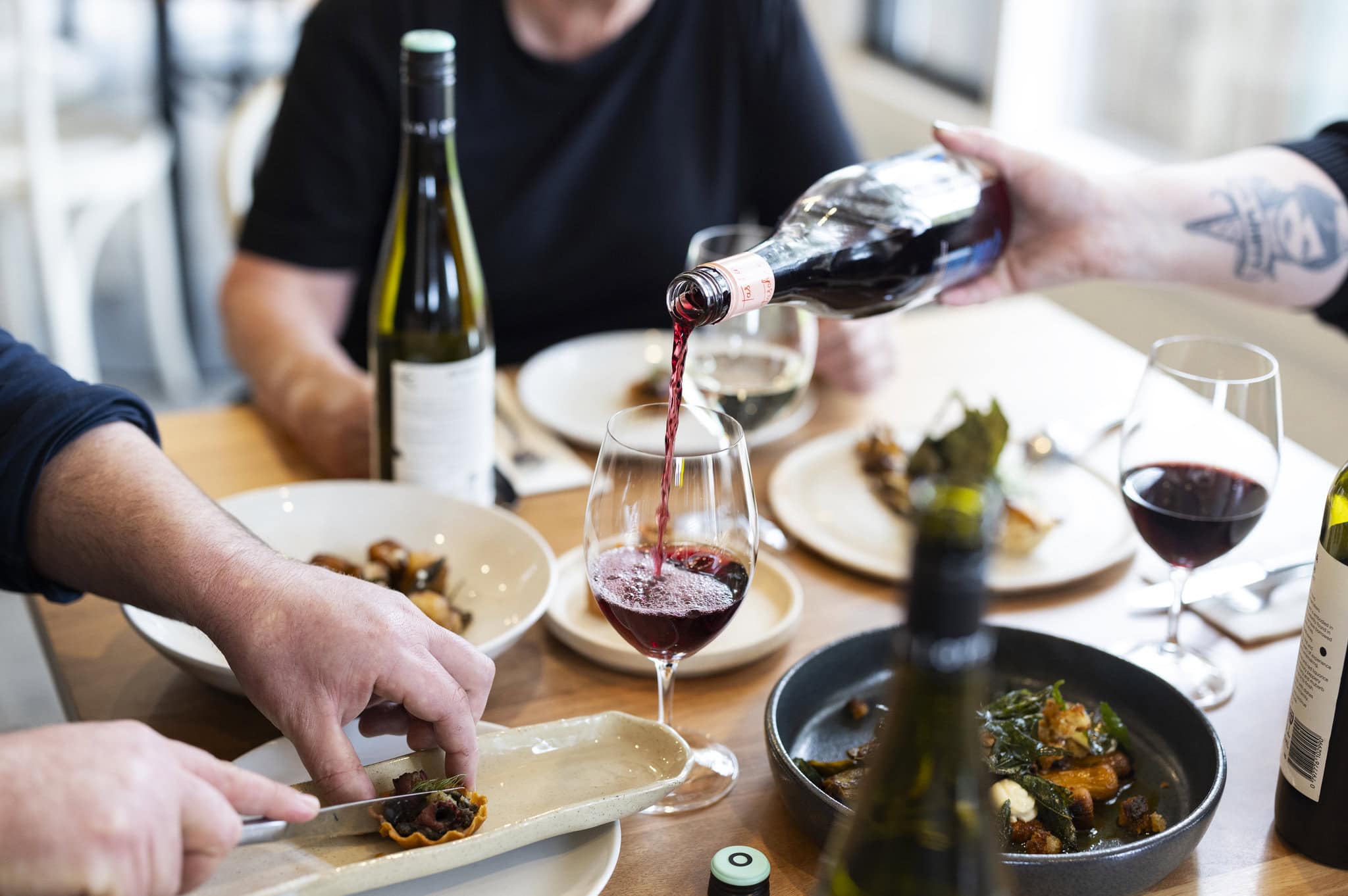 People dining at a restaurant table. Someone is pouring wine and another person is cutting some food.