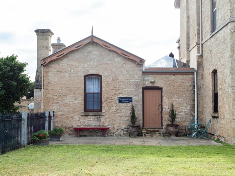 A photo of an old sandstone cottage, adjoining a large sandstone building (out of picture).