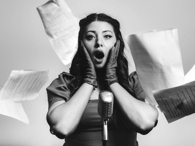 Black and white, actress stands in front of microphone gasping with hands on face, script pages fly