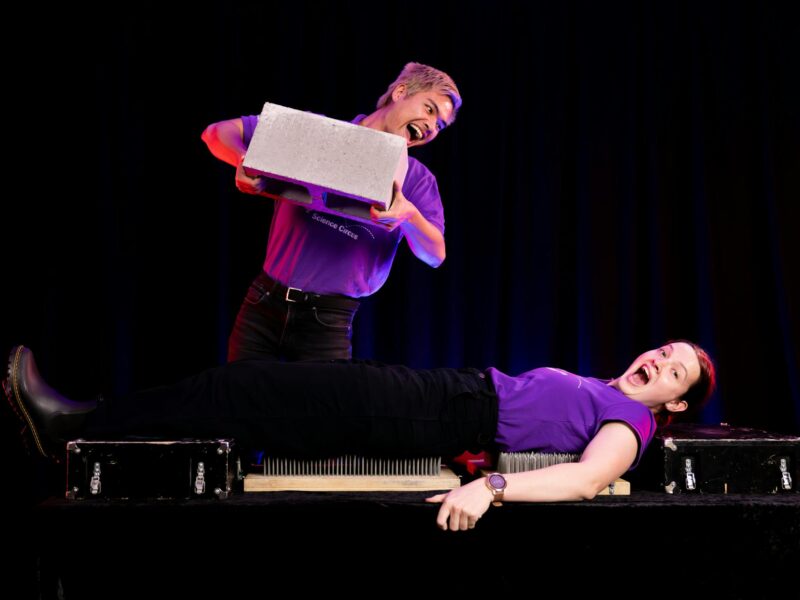 Two Questacon presenters perform a science demonstration involving laying on a bed of nails