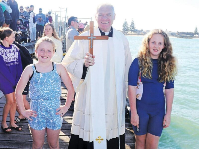 An older Anglican Priest with a clerical collar holding a wooden cross with two children.