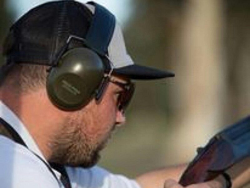 Man with a cap on and black earmuffs getting ready to line up and shoot a clay pigeon with a shotgun