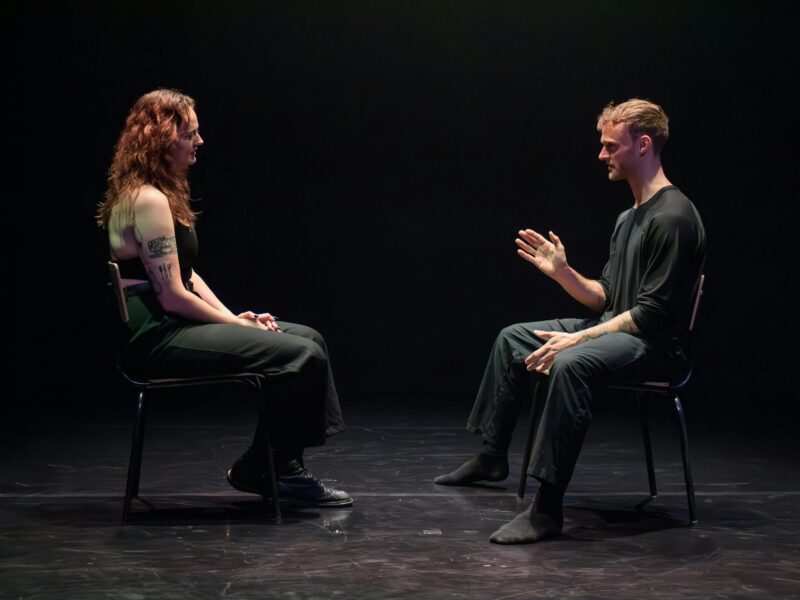 A man and woman sit in chairs facing one another while having a conversation on stage.