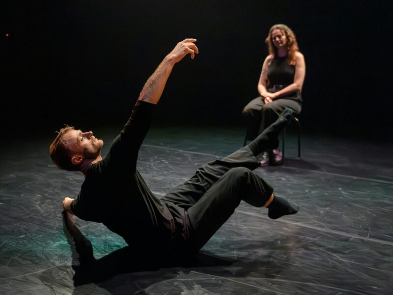 A man dressed in black dances on stage in front of a woman who sits in a chair watching.