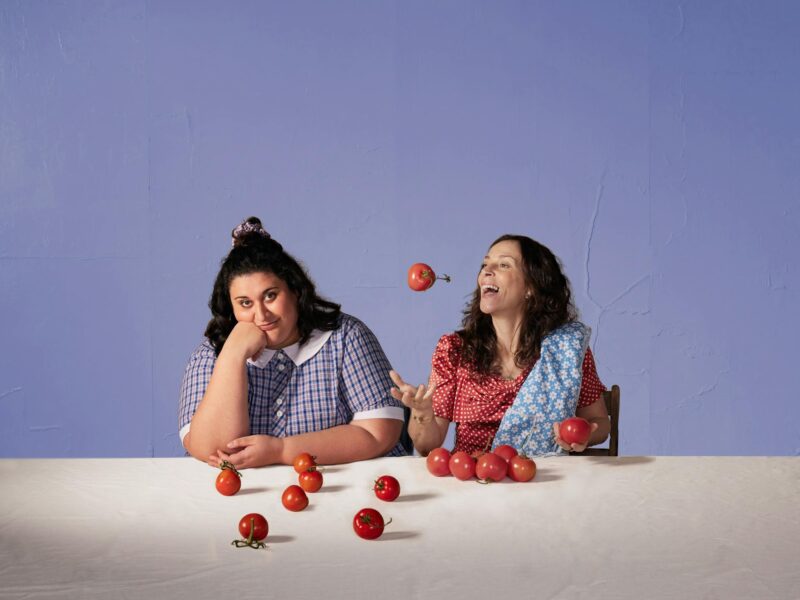 Two women sit at a table in front of many tomatoes, one woman joyfully juggles two of the tomatoes