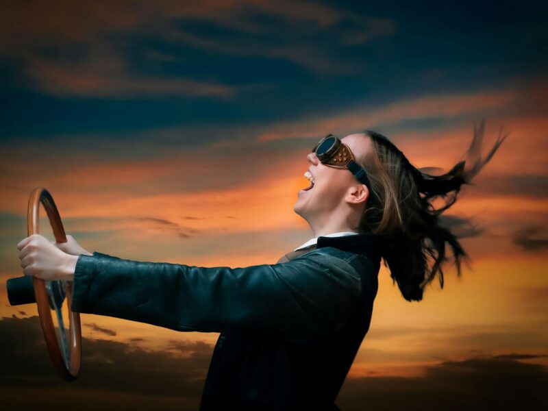A person holding a steering wheel and wearing goggles against a sunset backdrop pretending to drive.