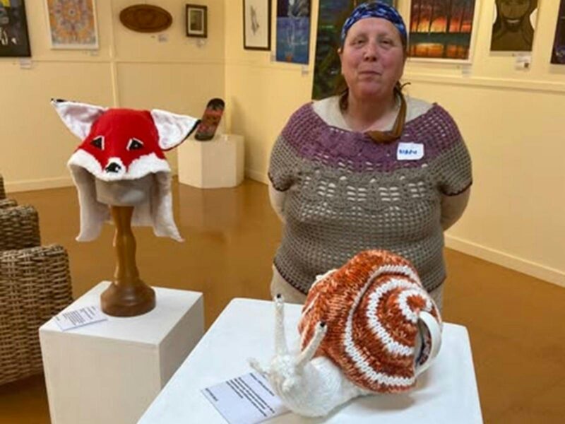 Mary posing in front of a red fox hat with white ears and a snail teapot cover that she made.