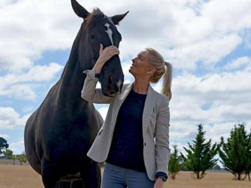 Liz Rymill patting a large black horse. Liz is wearing blue jeans, dark blue top and a grey jacket.