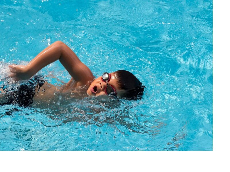 A young boy swimming in blue water. He has shorts on and a pair of goggles. Doing freestyle.