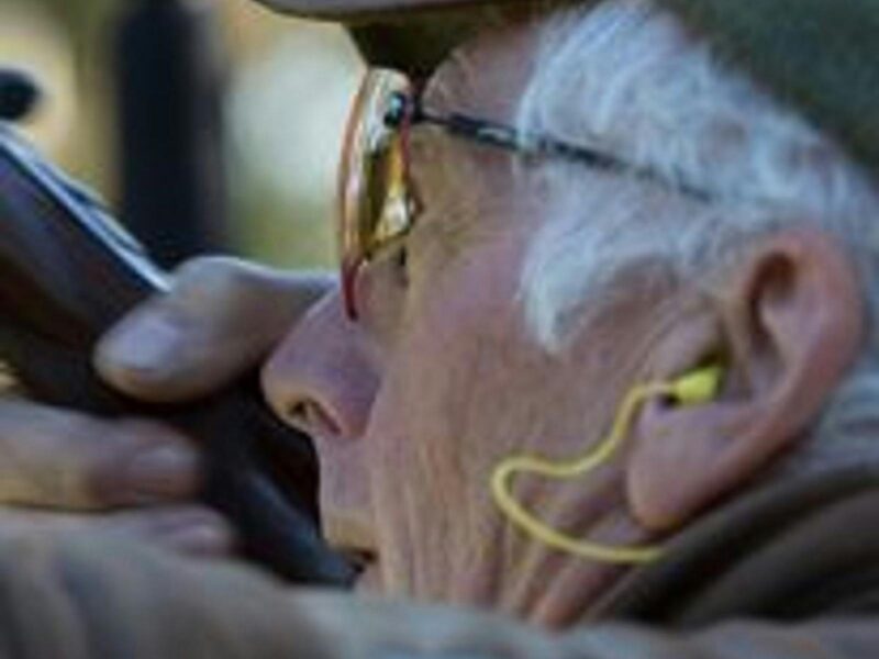 Elderly man with short grey hair and yellow earplugs poised to fire his shotgun at a clay pigeon.