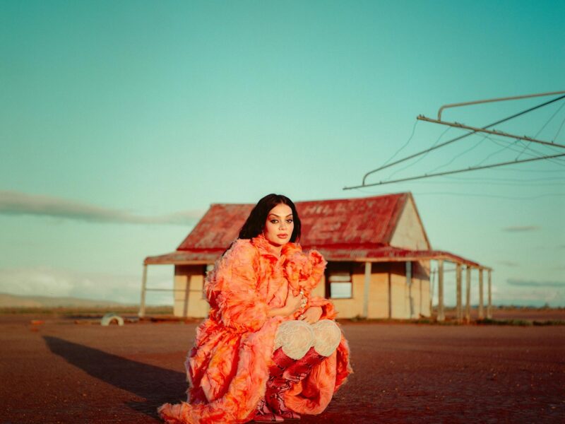 A lady with long back hair sitting in a rural setting wearing a long peach dress and high red boots