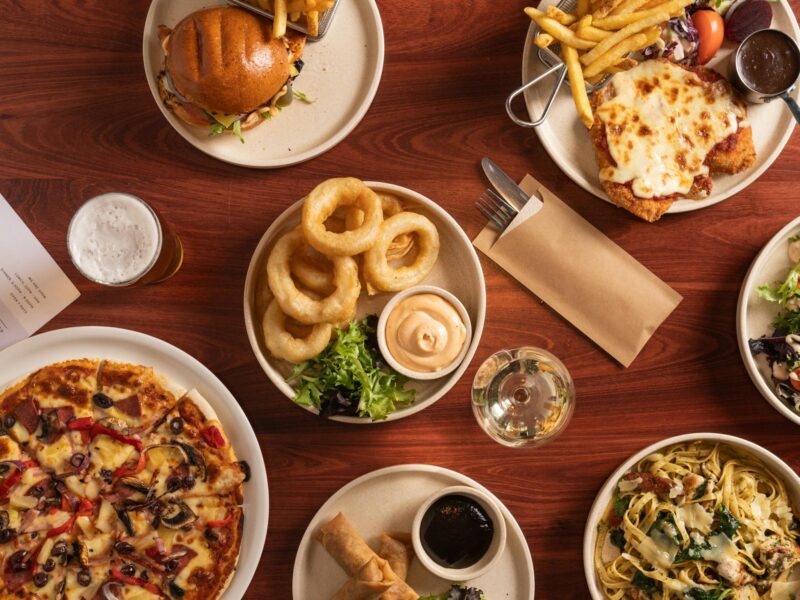 A table spread of pub meals, including onion rings, pizza, chicken parmigiana and a burger.