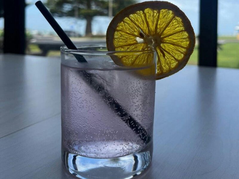 A delicious looking lilac drink in a glass tumbler with a black straw and  and orange slice on edge.