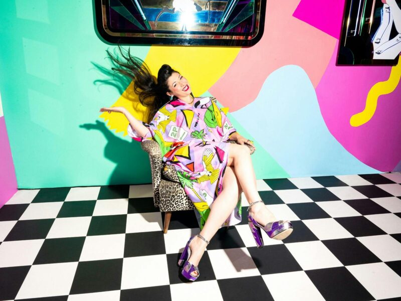 A woman flings her long dark hair while seated. The walls behind her are colourful as is her dress.