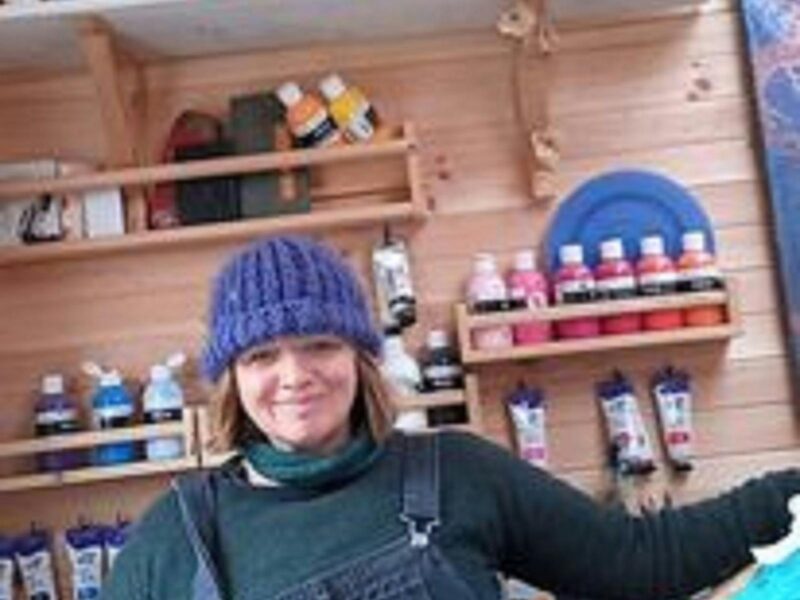 A photo of Selena Smith a talented artist, wearing a blue knitted hat blue, overalls  and green top.