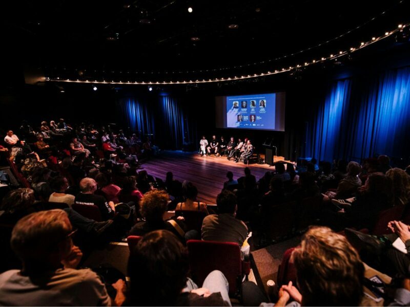A diverse crowd is seated, listening intently to a panel discussion taking place on stage.
