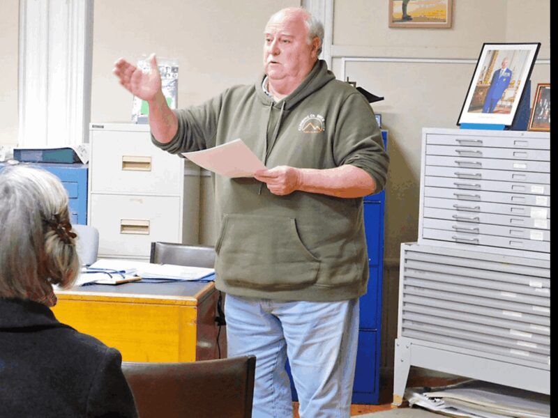 Photo of Steve Chappell with a green jumper with a hood. Holding some papers and his hand is forward