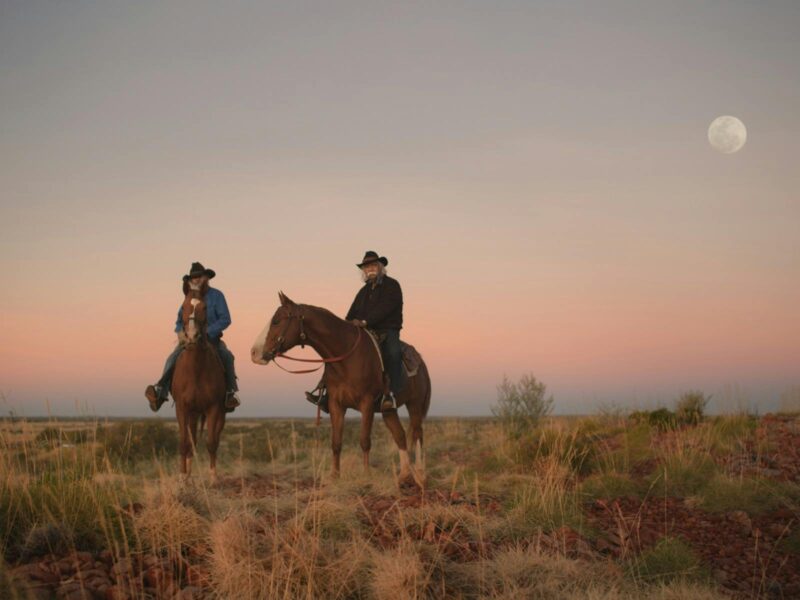 Two aboriginal men on horseback under a purple sky with a full moon.