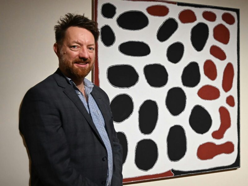A man with a navy suit and short spiky hair stands in front of an abstract painted piece of art