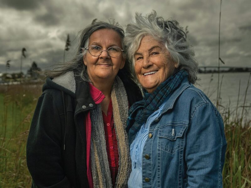 Two First Nations female Elders in front of water and a moody sky.
